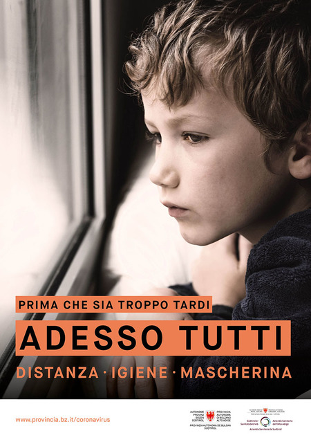 Un bambino triste guarda fuori dalla finestra. L’immagine è accompagnata dallo slogan: Prima che sia troppo tardi. Adesso tutti. Distanza, igiene, mascherina.
